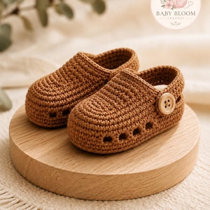 May include: A pair of brown crochet baby shoes featuring a rounded toe, perforated side details, and a single light-colored wooden button on the side strap. The shoes are textured with a visible yarn stitch pattern and have a slip-on design.