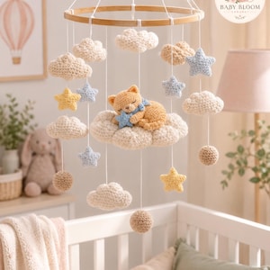 May include: A hanging nursery mobile featuring a circular wooden frame with white strings suspending crocheted elements including a sleeping orange cat, beige clouds, light blue and yellow stars, and small beige spheres in a soft color palette.