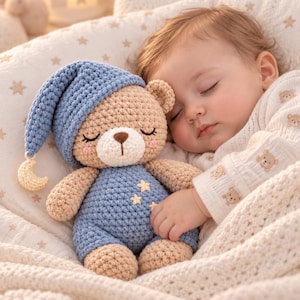 Puede incluir: Un oso de peluche tejido a ganchillo con cuerpo color canela, un gorro de dormir azul con una luna creciente amarilla colgante y un overol azul adornado con pequeñas estrellas amarillas. El oso tiene ojos cerrados, nariz marrón y mejillas rosadas.