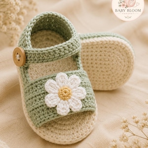 Op de afbeelding: Een paar gehaakte babysandalen in saliegroen en crème, voorzien van een witte madeliefjesapplicatie met een geel hartje op de voorste band, een houten knoopsluiting aan de zijkant en een crèmekleurige zool met textuur.