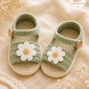 Peut inclure: Une paire de sandales pour b&eacute;b&eacute; en crochet vert sauge et cr&egrave;me, orn&eacute;es d'une marguerite blanche avec un c&oelig;ur jaune sur la bride avant, ferm&eacute;es par un petit bouton en bois sur le c&ocirc;t&eacute;, avec le logo Baby Bloom Crochet visible dans le coin.
