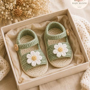 Peut inclure: Une paire de sandales pour b&eacute;b&eacute; en crochet vert sauge, orn&eacute;es d'une fleur marguerite blanche avec un c&oelig;ur jaune sur la bride avant. Chaque sandale poss&egrave;de un petit bouton en bois sur la bride lat&eacute;rale et une semelle cr&egrave;me, dans une bo&icirc;te.