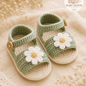 Peut inclure: Une paire de sandales pour b&eacute;b&eacute; en crochet vert sauge avec une applique marguerite blanche au centre jaune sur la bride avant et un petit bouton en bois sur la bride lat&eacute;rale. Le logo BABY BLOOM CROCHET est visible dans le coin sup&eacute;rieur droit.