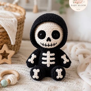 May include: A crocheted plush toy shaped like a skeleton wearing a black hooded outfit. The figure has a cream-colored skull face with large black eyes, a stitched mouth, and white bone-shaped accents on the chest, arms, and feet.