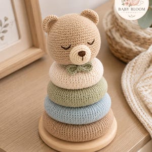 Puede incluir: Un juguete apilable de ganchillo con una cabeza de oso color canela con ojos cerrados y una pequeña nariz marrón, sobre cuatro anillos suaves y redondeados en color crema, verde salvia, azul claro y canela, sobre una base de madera.