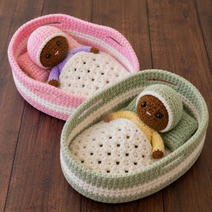 May include: Two crocheted dolls with dark brown skin and black eyes, each wearing a matching bonnet and resting in a small oval basket. One doll wears a purple shirt with a pink basket, while the other wears a yellow shirt with a green basket.