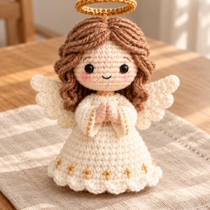 May include: A crocheted angel doll with light brown yarn hair, a gold halo, and white wings. The figure wears a cream-colored dress with gold embroidered cross details and has its hands pressed together in a prayer position.
