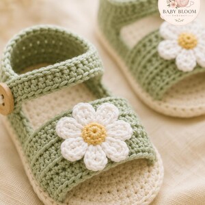 Peut inclure: Une paire de sandales pour b&eacute;b&eacute; en crochet vert sauge avec une fleur marguerite blanche au centre jaune sur la bride avant. Chaque sandale poss&egrave;de une semelle cr&egrave;me et un petit bouton en bois sur la bride lat&eacute;rale. Le texte Baby Bloom Crochet est visible.