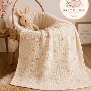 Può includere: Una coperta per neonati all'uncinetto color crema con un motivo a coste testurizzato e piccole applicazioni a forma di cuore rosa e beige distribuite uniformemente. La coperta è drappeggiata su una culla in vimini con un coniglio di peluche.