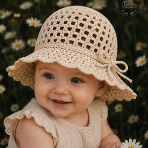 May include: A cream-colored crochet sun hat with a square-patterned crown and a scalloped brim featuring a side tie detail. The hat includes a circular logo in the upper right corner with the text Baby Bloom Crochet and a yarn ball icon.
