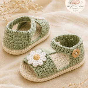 Peut inclure: Une paire de sandales au crochet vert sauge avec une fleur marguerite blanche appliqu&eacute;e sur le devant, une semelle cr&egrave;me et une fermeture &agrave; bouton en bois sur la bride de cheville. Les sandales portent le logo Baby Bloom Crochet.