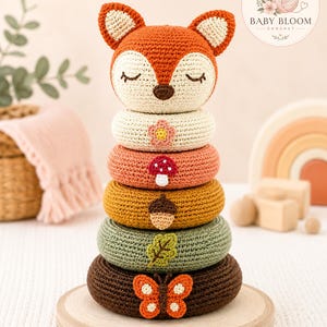 May include: A crochet stacking toy featuring a fox head topper and five rings in cream, peach, mustard, sage green, and dark brown. Each ring displays a unique applique: a flower, mushroom, acorn, leaf, and butterfly. The fox has closed eyes.