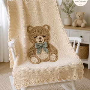 May include: A cream-colored crochet baby blanket featuring a scalloped edge and a large, centered applique of a brown teddy bear with closed eyes, a stitched nose, and a sage green bow tie. The blanket is draped over a white wooden rocking chair.