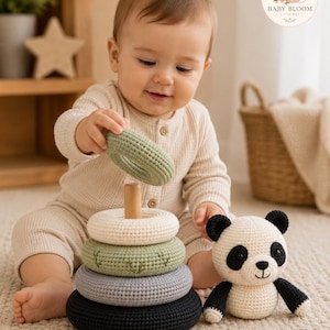 May include: A crochet stacking ring toy featuring four circular rings in sage green, cream, light green with leaf embroidery, and black, stacked on a wooden post. Beside the toy sits a small, round, cream and black crochet panda bear figure.
