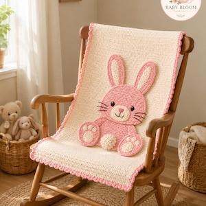 May include: A cream-coloured crochet blanket with a pink bunny appliqué featuring long ears, black eyes, and a white pom-pom tail. The blanket has a scalloped pink border and is draped over a wooden rocking chair. A logo reads BABY BLOOM CROCHET.