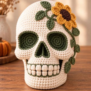 Puede incluir: Una calavera de ganchillo color crema con detalles de hilo verde oscuro en las cuencas de los ojos y la cavidad nasal. Un girasol amarillo con centro marrón oscuro y varias hojas y enredaderas de ganchillo verde oscuro adornan el lateral.