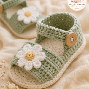 Peut inclure: Une paire de sandales pour b&eacute;b&eacute; en crochet vert sauge avec une application de marguerite blanche au centre jaune sur la bride avant. Chaque sandale poss&egrave;de une semelle cr&egrave;me et un bouton en bois rond sur la bride lat&eacute;rale. Le logo indique BABY BLOOM.