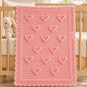 May include: A rectangular crochet blanket in a soft pink hue, featuring a repeating pattern of raised bobble stitch hearts across the center. The blanket is finished with a decorative scalloped edge border and a solid textured stitch base.