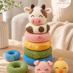 Puede incluir: Un juguete apilable de ganchillo con una cabeza de vaca en la parte superior, seguido de anillos en blanco con manchas marrones, rosa, amarillo, verde y azul. Dos anillos adicionales en azul y verde, una cabeza de cerdo rosa y un pollito amarillo.