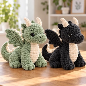 May include: Two crocheted dragon plush toys, one in sage green and one in black, both featuring cream-colored bellies, horns, and tail spikes. Each dragon has large dark eyes, textured yarn bodies, wings, and a seated posture with a curled tail.