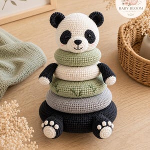 Puede incluir: Un juguete apilable de ganchillo con forma de panda, que incluye una cabeza de panda en la parte superior, dos brazos negros y dos pies negros con almohadillas blancas. La torre consta de cuatro anillos en verde salvia, crema, gris y negro.