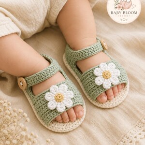 Peut inclure: Une paire de sandales au crochet vert sauge avec une fleur de marguerite blanche au centre jaune sur la bride avant. Les sandales ont une semelle cr&egrave;me et un petit bouton en bois sur la bride lat&eacute;rale. Un logo indique BABY BLOOM CROCHET.