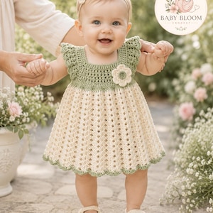 May include: A crochet dress featuring a sage green bodice with ruffled sleeves and a cream-coloured skirt with a textured vertical pattern. The dress is accented with a single cream flower appliqué at the chest and a scalloped sage green hemline.