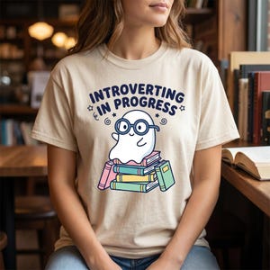 May include: A beige short-sleeve t-shirt featuring a graphic of a white ghost wearing black glasses, sitting on a stack of colorful books. The text above the ghost reads "INTROVERTING IN PROGRESS" in dark blue capital letters with small star accents.