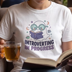 May include: A white t-shirt featuring a graphic of a ghost wearing round glasses and reading a book titled Stories. The design includes small stars, moons, and two mugs of tea, with the text Introverting In Progress printed in dark purple letters.