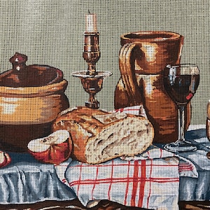May include: A needlepoint canvas depicting a still life with a brown lidded pot, a brass candlestick, a brown pitcher, a glass of red wine, a loaf of bread, a sliced apple, and a soft-boiled egg in a cup, all set on a blue and white checkered cloth.