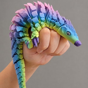 Könnte beinhalten: Ein 3D-gedrucktes, bewegliches Pangolin-Spielzeug mit einem lebendigen Regenbogen-Farbverlauf, der von Violett zu Blau, Grün und Gelb übergeht. Die Figur hat einen segmentierten, flexiblen Körper mit überlappenden schuppenartigen Platten und einer spitzen Schnauze.