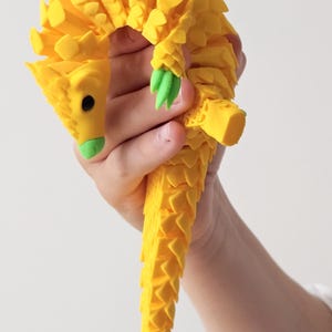 Könnte beinhalten: Eine gelbe, bewegliche Pangolin-Figur mit einer strukturierten, schuppenartigen Oberfläche, einem leuchtend grünen Schnauzenbereich und grünen Krallen an den Füßen. Das Tier hat einen langen, segmentierten Schwanz und ein kleines schwarzes Auge.