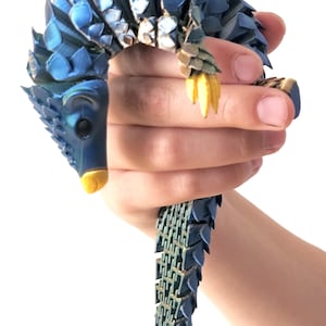 Op de afbeelding: Een blauw gearticuleerd pangolin-figuurtje met een gelaagde, schubachtige textuur over het hele lichaam. Het wezen heeft een gebogen rug, een puntige kop met een gele snuit en gele klauwen aan de voorpoot. Het oppervlak heeft een metaalachtige glans.