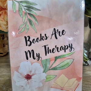 May include: A rectangular card featuring a soft peach watercolor background with thin wavy lines. The text "Books Are My Therapy" is printed in black script. The design includes a white flower with a red center, green leaves, and an open book.
