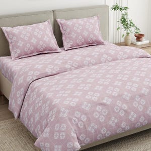 Puede incluir: Un juego de cama rosa claro con un patrón geométrico blanco repetitivo de formas florales de cuatro pétalos dispuestas en cuadrícula. El conjunto incluye una funda nórdica y dos fundas de almohada rectangulares a juego.