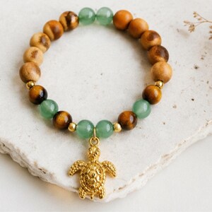 Peut inclure: Un bracelet de perles composé d'une alternance de perles en bois brun, de perles œil-de-tigre et de perles lisses vert jade. Le bracelet comprend de petites entretoises en métal doré et une breloque centrale en forme de tortue de mer.