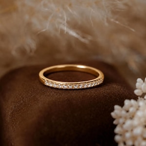 Peut inclure: Une fine bague en or ornée d'une rangée de petites pierres claires taillées en rond, incrustées sur la moitié avant de la surface métallique. La bague présente une finition polie et un design circulaire simple d'une largeur constante.