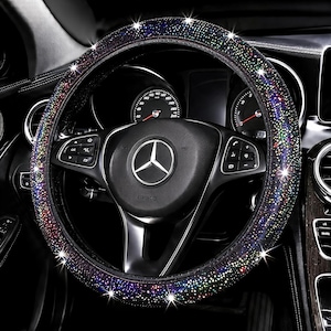 May include: A black steering wheel cover adorned with numerous small, reflective, multi-colored rhinestones that create a sparkling effect. The cover is fitted onto a black steering wheel featuring a central silver Mercedes-Benz logo emblem.
