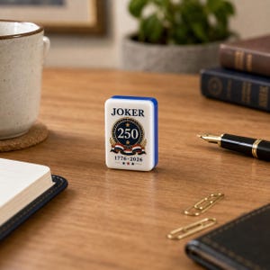 May include: A white rectangular mahjong tile with blue edges featuring the text JOKER at the top, a central circular emblem with the number 250, laurel branches, a red, white, and blue ribbon, and the dates 1776-2026 with three small stars below.