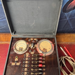 May include: A vintage electrical testing device housed in a dark wooden box with a hinged lid. The panel features two circular analog gauges labeled Volts and Milliamperes, multiple metal input ports, and a series of red and black toggle switches.