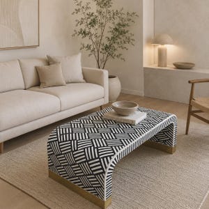May include: A low-profile rectangular coffee table featuring a bold black and white geometric woven pattern across its surface and sides. The table has a curved waterfall design with metallic gold-toned base accents at both ends.