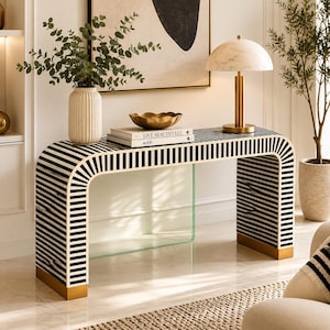Puede incluir: Una mesa consola con un marco curvo estilo cascada que presenta un patrón de rayas blancas y negras y detalles dorados en la base. La superficie de la mesa sostiene un jarrón blanco acanalado con vegetación, un cuenco dorado y un libro titulado Live Beautifully.