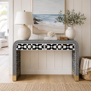 May include: A black and white console table with a geometric pattern, two drawers, and gold-toned base accents. The table features a curved frame design and is topped with a white lamp, a coral sculpture, two stacked books, and a vase with greenery.