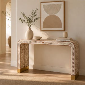 May include: A rectangular console table with rounded corners, featuring an intricate white and brown leaf-patterned inlay. The table has gold-toned metal feet and is topped with a white vase containing greenery, a small bowl, and two stacked books.