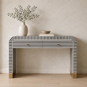 Könnte beinhalten: Ein Konsolentisch mit abgerundeter Oberseite und zwei Schubladen, der ein schwarz-weißes Streifenmuster aufweist. Der Tisch hat goldfarbene Metallfüße und zwei kleine runde Knöpfe. Eine beige Vase mit grünem Blattwerk und eine Schale stehen darauf.
