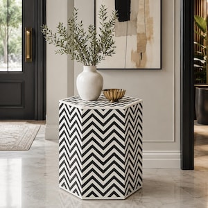 May include: A hexagonal side table featuring a black and white chevron pattern. On top of the table sits a white ceramic vase filled with green leafy branches and a small, fluted gold-toned metal bowl. The table has a flat top and six sides.