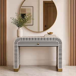 May include: A console table featuring a black and white striped pattern with a curved frame and a single drawer with a small gold knob. The base of the table legs is finished in gold. The table is topped with a white ribbed vase and a small bowl.