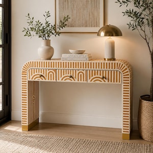May include: A rectangular console table featuring a white and mustard yellow striped pattern with arched designs on the three front drawers. The table has a curved frame and gold-toned metal feet at the base of each side, providing a stable foundation.
