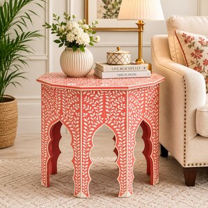 May include: An octagonal side table featuring a vibrant coral-red finish with intricate white floral vine inlays. The table has four arched legs and a flat top surface. It is decorated with a white ribbed vase of flowers and two stacked books.