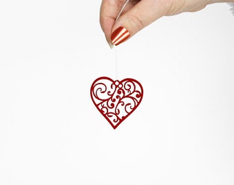 May include: A red heart-shaped ornament with a delicate, intricate design. The ornament is hanging from a string held by a hand.